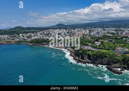 Luftaufnahme des Jeongbang-Wasserfalls, der über Küstenklippen ins Meer stürzt, mit der Stadt Seogwipo und üppiger Vegetation im Hintergrund in Südkorea Stockfoto