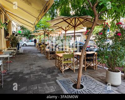 Außenbestuhlung eines Cafés unter beigen Sonnenschirmen entlang einer gepflasterten Straße. Bäume mit grünen Blättern und rosa Blüten in der Nähe. Holzstühle und Tische sind bereitgestellt für Stockfoto
