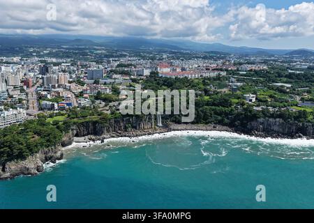 Luftaufnahme des Jeongbang-Wasserfalls, der über Küstenklippen in das türkisfarbene Meer stürzt, mit der städtischen Skyline von Seogwipo, Jeju Island, Südkorea. Stockfoto