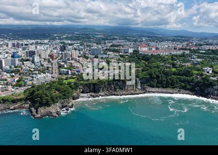 Luftaufnahme des Jeongbang-Wasserfalls, der über felsige Klippen ins Meer stürzt, umgeben von üppigen grünen Bäumen und der städtischen Skyline von Seogwipo im b Stockfoto