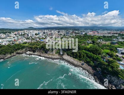 Luftaufnahme des Jeongbang-Wasserfalls, der über felsige Klippen ins Meer stürzt, umgeben von üppigen grünen Bäumen und der städtischen Skyline von Seogwipo, Jeju Island Stockfoto