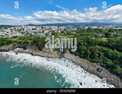 Luftbild des Jeongbang-Wasserfalls, der über Basaltklippen ins Meer stürzt, mit der städtischen Skyline von Seogwipo im Hintergrund auf Jeju Island, Südkorea Stockfoto