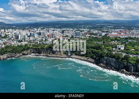 Luftaufnahme des Jeongbang-Wasserfalls, der über Küstenklippen in den Ozean stürzt, mit der Stadtlandschaft von Seogwipo im Hintergrund, Südkorea. Stockfoto