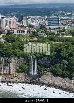Luftaufnahme des Jeongbang-Wasserfalls, der über Basaltklippen in den Ozean stürzt, mit der Stadt Seogwipo und üppiger Vegetation im Hintergrund in Sou Stockfoto