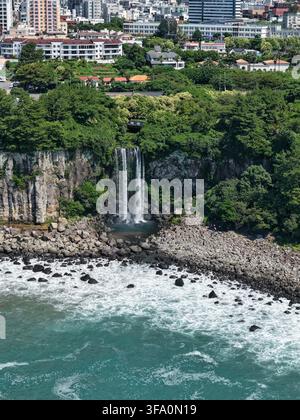Luftaufnahme des Jeongbang-Wasserfalls, der über felsige Klippen ins Meer stürzt, umgeben von üppiger grüner Vegetation und städtischen Gebäuden in Südkorea. Stockfoto