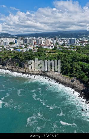 Luftaufnahme des Jeongbang-Wasserfalls, der über eine felsige Klippe ins Meer stürzt, mit üppiger Vegetation und der Stadt Skyline im Hintergrund in Südkorea. Stockfoto