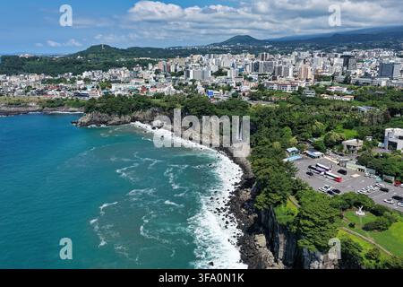 Luftbild des Jeongbang-Wasserfalls, der über Küstenklippen ins Meer stürzt, mit der Stadt Seogwipo und üppiger Vegetation in Seogwipo, Jeju Island, So Stockfoto