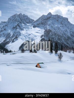Blick auf eine kleine Holzhütte, die in einem weiten schneebedeckten Tal liegt, umgeben von hohen schneebedeckten Bergen und Kiefern in Kashmir, Himachal Pr Stockfoto