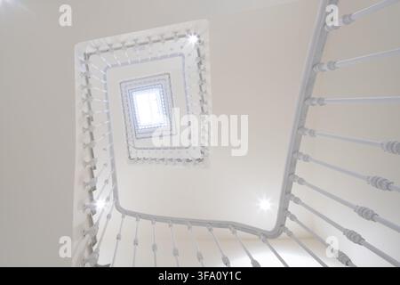 Aufblick durch eine helle neoklassizistische Bürotreppe, mit weißen Geländern, kunstvollen Brüstungen, geometrischen Ebenen und einem leuchtenden Oberlicht Stockfoto