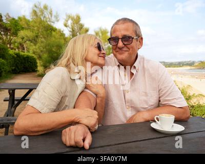 Lächelndes Seniorenpaar teilt einen zarten Moment an einem Strandcafé-Tisch, genießt Kaffee und die Gesellschaft des anderen während eines entspannten Küstenlebensstils hol Stockfoto