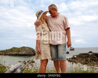 Glückliches Seniorenpaar umarmt sich und lächelt sich auf einem felsigen Strand während eines Seebadurlaubs an, wobei die Frau einen Koffer unter einer bewölkten Sonne hält Stockfoto