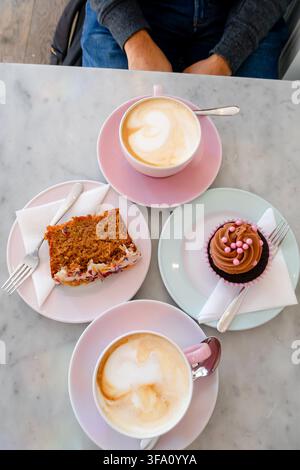 Oberblick auf einen Cafétisch mit zwei Tassen Latte, einer Scheibe mehrschichtigem Kuchen und einem glasierten Cupcake auf pastellfarbenen Tellern, wobei eine sitzende Person sichtbar ist Stockfoto