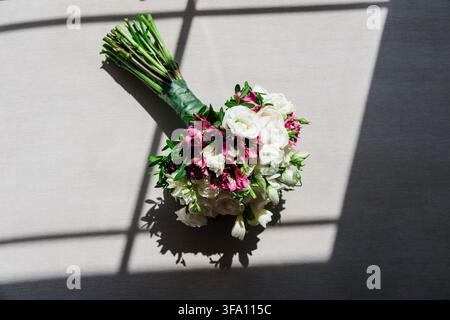 Eleganter Brautstrauß aus weißen Rosen und pinken Blumen ruht auf einem grauen Boden, beleuchtet von Fensterlicht und dramatischen Schatten für eine romantische Hochzeit Stockfoto