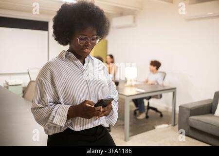 Schwarzer Profi mit Brille nutzt ein Smartphone in einem hellen modernen Coworking-Büro, während Kollegen im Hintergrund an Schreibtischen arbeiten. Stockfoto