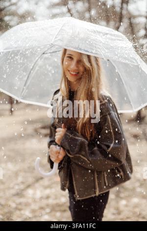 Lächelndes Mädchen steht in einem Park während des Regens und hält einen transparenten Regenschirm, während Regentropfen um sie herum fallen. Sie trägt eine dunkle Jacke an einem kühlen Herbsttag Stockfoto