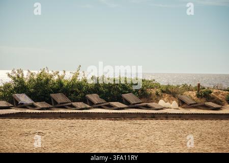 Leere hölzerne Sonnenliegen säumen einen sandigen Sommerstrand in Saulkrasti, Lettland, mit Blick auf die ruhige Ostsee unter einem blassblauen Himmel, mit Küstenstrauch Stockfoto
