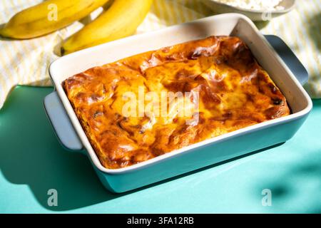 Hausgemachte proteinreiche Hüttenkäse-Auflauf mit einer goldenen gebackenen Kruste in einer Keramikschale, serviert auf einem türkisfarbenen Tisch mit reifer Banane für eine wholeso Stockfoto