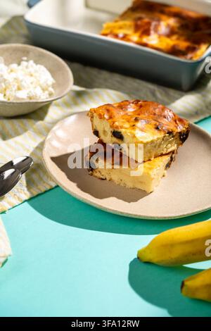 Goldene Hüttenkäse-Backscheiben mit Rosinen werden auf einem Teller neben frischem Hüttenkäse, einer Backform und Bananen für eine nahrhafte Hoch-Pro-Portion serviert Stockfoto