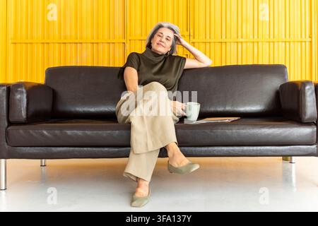 Reife grauhaarige Frau sitzt bequem auf einem Ledersofa mit einer Kaffeetasse und einem Ordner in der Nähe, eine Hand auf ihrem Kopf ruhend während einer entspannten informellen Stockfoto