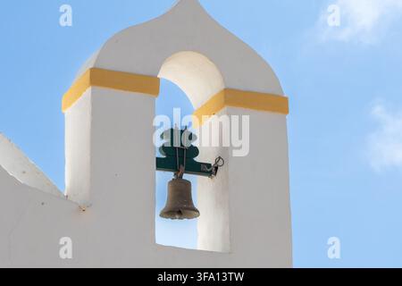 Eine Nahaufnahme eines weißen Glockenturms mit einer Bronzeglocke, die an einer grünen Holzkonstruktion hängt, vor einem klaren blauen Himmel mit vereinzelten Wolken Stockfoto