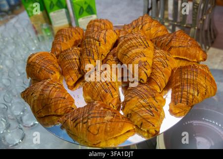 Frisch gebackene goldene Buttercroissants, die mit reichhaltiger Schokoladenglasur beträufelt sind, wunderschön auf einem verspiegelten Tablett bei einem Buffet serviert. Perfektes, köstliches Gebäck Stockfoto
