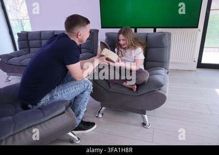 Lächelnder Mann gibt einer freudigen Frau, die auf einem grauen Stuhl in einem modernen Wohnzimmer sitzt, eine geöffnete Geschenkschachtel mit leerem grünen Bildschirm, hellem Tageslicht, lässig Stockfoto