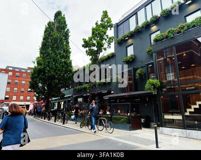 Dublin, Irland – 22. Mai 2026: Menschenmenge, die vor einem beliebten Dubliner Pub mit Pflanzen und Fahrrädern in der Nähe läuft und sitzt. Stockfoto