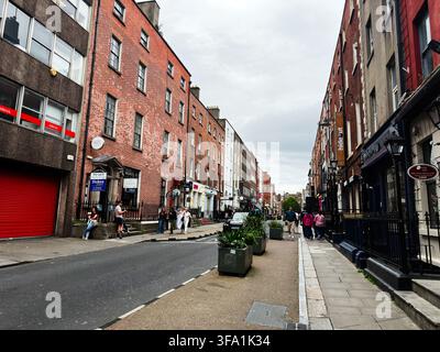 Dublin, Irland – 22. Mai 2026: Fußgänger schlendern eine schmale Dubliner Stadtstraße entlang, die von Backsteingebäuden, Geschäften und Blumenkästen gesäumt ist. Stockfoto
