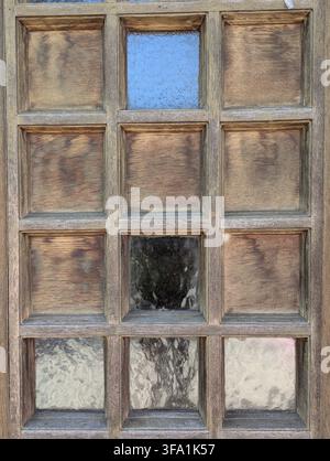 Nahaufnahme eines verwitterten hölzernen Fensterrahmens, der mattierte Glasscheiben trennt, mit sichtbar gealterter Maserung und gedämpften Farben, durch eine Scheibe eine kühle blaue Tönung. Tex Stockfoto
