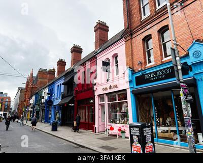 Dublin, Irland – 22. Mai 2026: Lebendige Dubliner Straße mit farbenfrohen Schaufenstern, Einkäufern, Backsteinarchitektur und einem stimmungsvollen bewölkten Himmel. Stockfoto