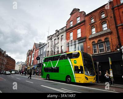 Dublin, Irland – 22. Mai 2026: Ein grüner und gelber Doppeldeckerbus steht auf einer Dubliner Straße, die von historischen Backsteinläden und Fußgängern gesäumt ist. Stockfoto