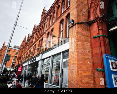 Dublin, Irland – 22. Mai 2026: Belebte Dubliner Straße mit roten Backsteinfassaden aus der viktorianischen Ära, Geschäften und Fußgängern im städtischen Stadtzentrum. Stockfoto