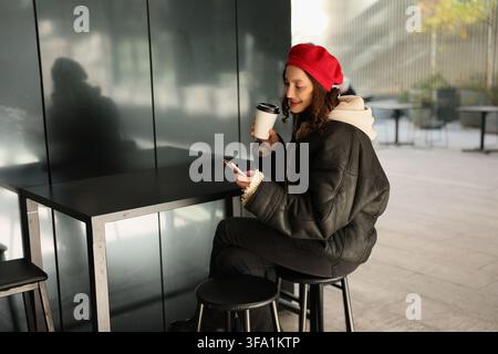 Junge Frau mit rotem Barett sitzt an modernem Cafétisch, lächelt auf ihr Smartphone, während sie einen To-go-Kaffeebecher hält, trägt dunklen Mantel und Schal daneben Stockfoto