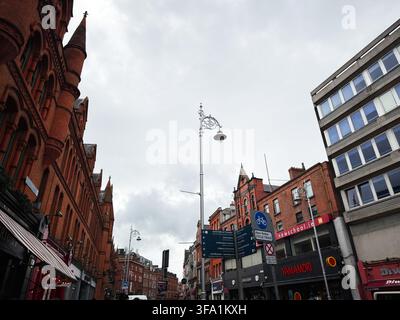 Dublin, Irland – 22. Mai 2026: Urbane Straßenszene mit roten Backstein-Viktorianischen Gebäuden, bewölktem Himmel, Geschäften und Laternenpfählen in Dublin. Stockfoto
