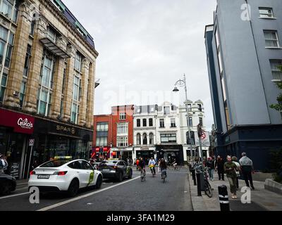 Dublin, Irland - 22. Mai 2026: Fußgänger und Radfahrer navigieren durch eine belebte Dubliner Stadtstraße, die von historischen Gebäuden und Geschäften gesäumt ist. Stockfoto