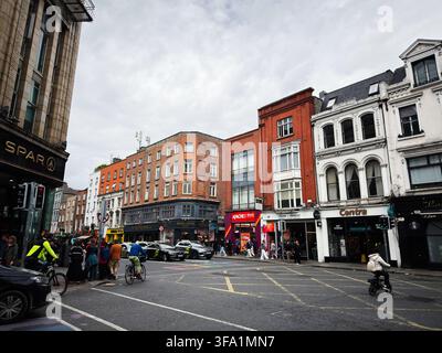 Dublin, Irland - 22. Mai 2026: Fußgänger und Radfahrer navigieren durch eine belebte Dubliner Straße, die von Geschäften, Taxis und viktorianischen und modernen Gebäuden gesäumt ist. Stockfoto