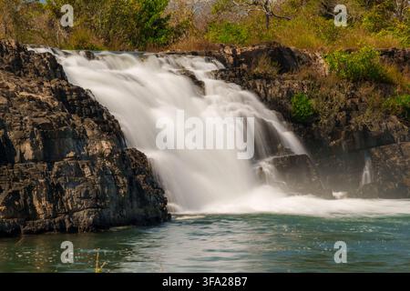 Ein mächtiger Abschnitt des Tad-Faek-Wasserfalls stürzt über dunkles, gegliedertes Gestein in ein klares Flussbecken, umgeben von Sprühnebel und Vegetation. Stockfoto