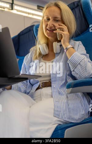 Reife Geschäftsfrau telefoniert mit Kopfhörern, die um ihren Hals hängen, während sie im Zug sitzt. Konzept von professioneller Kommunikation und Busi Stockfoto