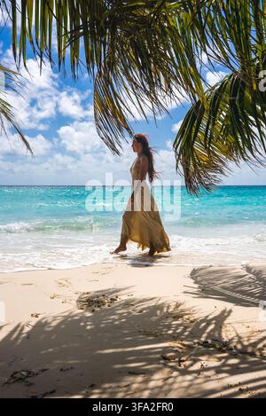 Eine Frau in einem langen Kleid geht barfuß entlang der sandigen Meeresküste unter tropischen Palmen im Tageslicht auf den Malediven. Stockfoto
