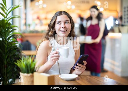Frau, die das Handy in der Hand hält und Kaffee trinkt, während sie an einem Tisch sitzt Stockfoto