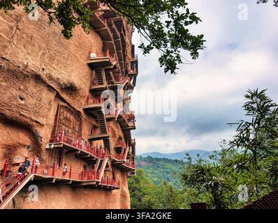 Ansicht der historischen Maijishan-Höhlen mit kunstvollen hölzernen Stegen, die in die steile rote Sandsteinklippenwand gehauen sind, mit üppigem grünen Wald in T Stockfoto