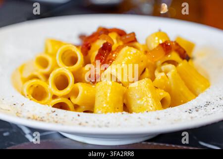 Eine Nahaufnahme einer herzhaften Portion Rigatoni-Pasta, die in einer reichhaltigen, goldenen Sauce überzogen und mit herzhaften Pancetta-Stücken belegt ist, bestäubt mit geriebenem Käse Stockfoto