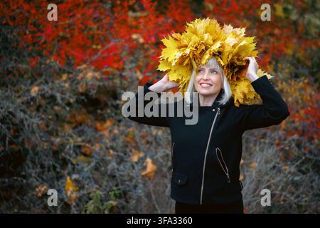 Eine lächelnde blonde Frau in einer schwarzen Jacke posiert in einem Herbstpark. Sie trägt eine große Krone aus hellgelben Ahornblättern, vor einem verschwommenen Stockfoto