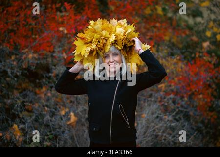 Ein Porträt einer fröhlichen blonden Frau in einer schwarzen Jacke, die in die Kamera schaut. Sie trägt eine üppige Krone aus gelben Ahornblättern auf dem Kopf, mit einem unscharfen Stockfoto