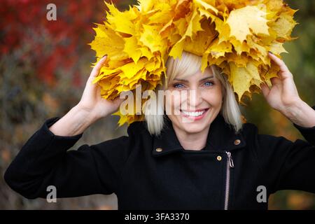 Ein Nahaufnahme-Porträt einer fröhlichen blonden Frau, die einen lebendigen Kranz aus gelben Ahornblättern auf dem Kopf hält. Sie trägt eine schwarze Jacke und lächelt warmherzig a Stockfoto