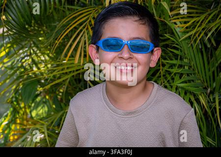 Junge mit blauer Sonnenbrille und gemustertem Hemd, der selbstbewusst lächelt, während er vor üppigen grünen tropischen Palmen steht, und eine helle s Stockfoto