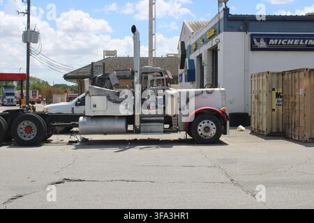 Schwerlastwagen vor einer Autowerkstatt geparkt. Metallrahmen zeigt Anzeichen von Abnutzung. Rote Räder kontrastieren mit verblasster weißer Fahrerkabine. Arbeiterwerkzeuge und c Stockfoto