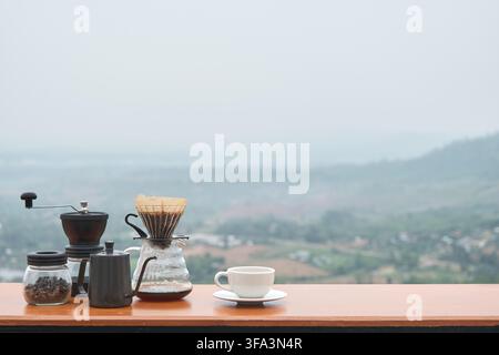 Tropfender Kaffee mit Mühle, Glas und Kaffeebohnen auf dem Tisch in der Landschaft mit Bergblick am Morgen mit verschwommenem, verblasstem Nebelhintergrund Stockfoto