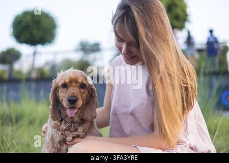 Eine Frau lächelt und streichelt ihren Hund Cocker Spaniel, während sie auf dem Gras sitzt Stockfoto