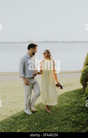 Ein Paar geht Hand in Hand am Seeufer entlang, lächelt und genießt die warme Sonne. Die friedliche Landschaft trägt zu ihrem freudigen Moment bei. Stockfoto